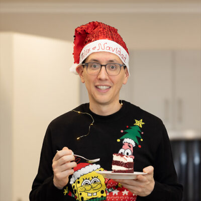 A man wearing a festive Christmas sweater, smiling and standing against a holiday-themed background.