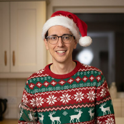 A man wearing a Christmas sweater is holding a knife, smiling in a festive kitchen setting.